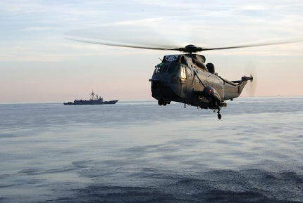 A Canadian Forces CH-124 Sea King helicopter hovers above the sea off the coast of Greece. The USS Boone is in the background. The helicopter is attached to the Canadian destroyer HMCS IROQUOIS
